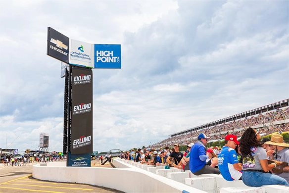 Race fans seated at Pocono Raceway near a tall sign featuring Eklund Farm Machinery branding and event sponsors.
