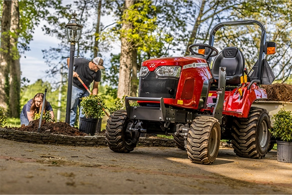 Massey Ferguson GC1700 Series subcompact tractor parked in a residential landscape setting, with two people working in the background, showcasing its versatility for yard work, gardening, and light-duty tasks.