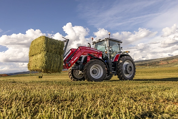 Massey Ferguson 8S tractor lifting a large square hay bale in a wide open field under blue skies