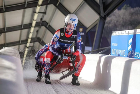 Team USA luge athletes racing in a doubles event on an ice track during international competition.